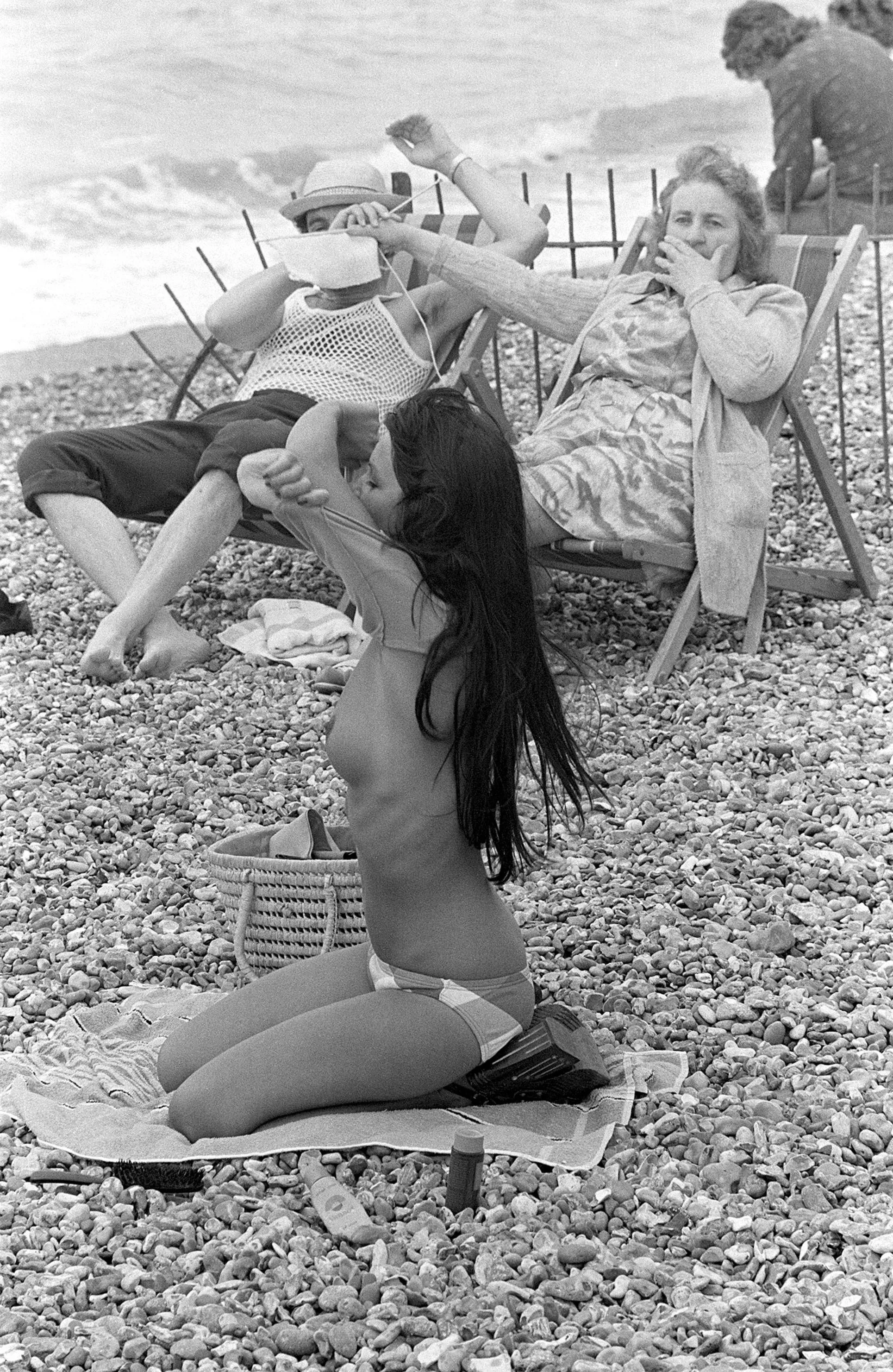 Woman covering man's eyes with her knitting at sight of young woman taking off her top on the beach in France, 1974 Woman covering man's eyes with her knitting at sight of young woman taking off her top on the beach in France, 1974
